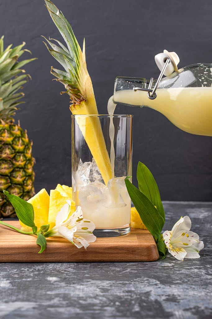 Delicious pineapple juice being poured into a glass with fresh slices and flowers on a wooden board.